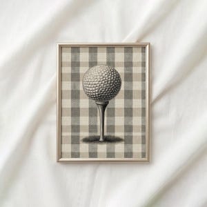 May include: Framed golf-themed art print. The artwork features a golf ball on a tee, set against a neutral-toned plaid background. The print is encased in a silver-colored frame, offering a classic and understated aesthetic.