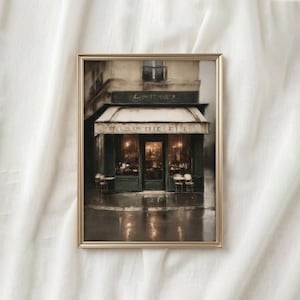 May include: Framed print of a Parisian cafe scene. The artwork depicts a building with a green awning and the text "L'ATELIER" above the entrance. The cafe's interior is illuminated, and the street reflects the lights.