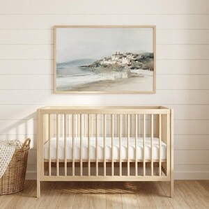 Puede incluir: Una cuna de madera clara con un colch&oacute;n blanco est&aacute; frente a una pared con una pintura de paisaje enmarcada. Una cesta tejida con una manta beige est&aacute; a la izquierda. La pintura representa una escena costera con un pueblo.