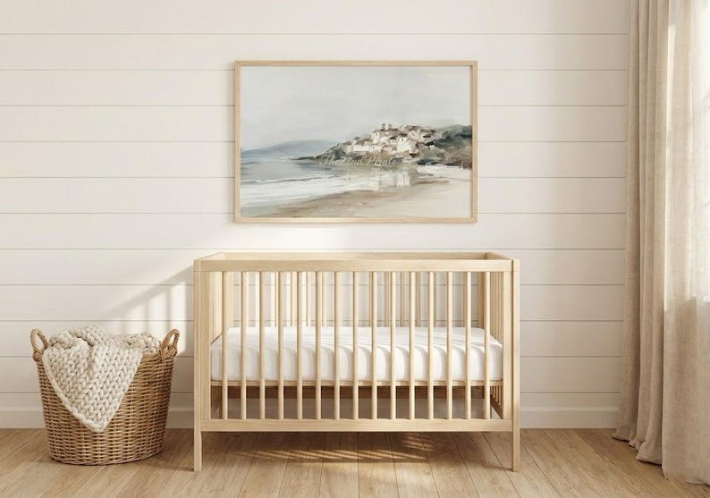 Puede incluir: Una cuna de madera clara con un colch&oacute;n blanco est&aacute; frente a una pared con una pintura de paisaje enmarcada. Una cesta tejida con una manta beige est&aacute; a la izquierda. La pintura representa una escena costera con un pueblo.