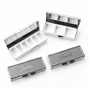 Puede incluir: Dos pastilleros rectangulares de metal plateado con divisores de plástico blanco en el interior, uno con seis compartimentos y otro con tres. Dos estuches cerrados de metal plateado tienen grabados los nombres Sophia y Daniel en letra cursiva.