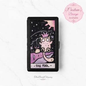 May include: A rectangular black case featuring a tarot card illustration titled The Fool. The artwork depicts a pale pink cat wearing a crown, sitting on a purple cliff under a dark starry sky with a sun, surrounded by small flowers.