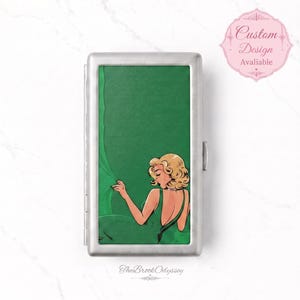 May include: A rectangular silver metal cigarette case featuring a green graphic illustration of a blonde person in a green dress reaching for a green curtain. The case has a hinged lid and a smooth metallic finish. The text The Brook Odyssey is printed at the bottom.