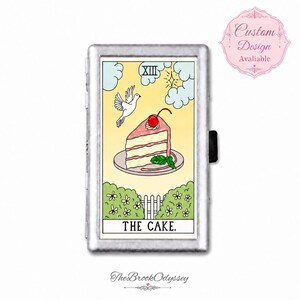 May include: A rectangular silver metal cigarette case featuring a tarot card illustration titled THE CAKE. The design shows a slice of cake with a cherry, a flying white dove, clouds, and a garden fence against a pale yellow background.