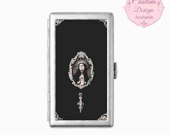 May include: A rectangular silver-toned metal cigarette case featuring a black front panel. The center displays an oval portrait of a figure in a habit within an ornate frame, with a decorative filigree element below and corner accents.
