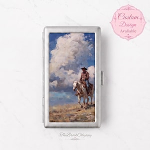 May include: A rectangular silver metal cigarette case featuring a printed oil painting of a cowboy riding a white horse across a dry landscape under a large, dramatic blue sky with white clouds. The case has a hinged side and a metallic finish.