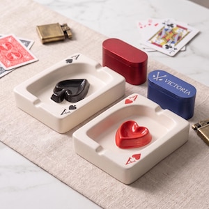 May include: Two rectangular off-white ceramic ashtrays shaped like playing cards. One features a black spade symbol in the center with matching spade icons at the corners, while the other has a red heart symbol with heart icons at the corners. Includes a red metal tin and a blue metal tin labeled Victoria.