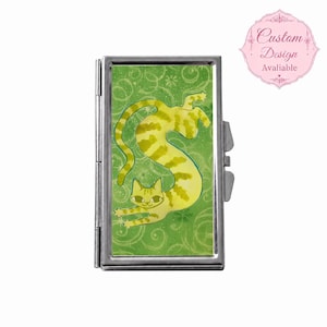 Könnte beinhalten: Ein rechteckiges Etui aus silbernem Metall mit grünem Hintergrund, Wirbelmuster und einer Illustration einer gelben getigerten Katze. Die Katze ist in einer langen, gestreckten Pose dargestellt. Ein rosa kreisförmiges Abzeichen zeigt Custom Design Avaliable.