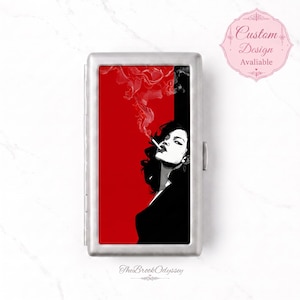 May include: A rectangular silver metal cigarette case featuring a graphic illustration of a person with dark hair smoking a cigarette. The background is split between solid red and black, with white smoke swirling above the figure's head.