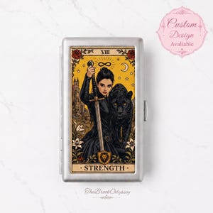 May include: A rectangular silver metal cigarette case featuring a Strength tarot card illustration. The design depicts a person in black clothing holding a sword next to a black panther, with a yellow background, roses, and the text STRENGTH.