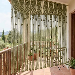 May include: An olive green macrame curtain featuring a series of repeating diamond-shaped knots along the top section, transitioning into long, vertical hanging cords. The curtain is suspended from a wooden rod and displays the ARVORA logo in the corner.