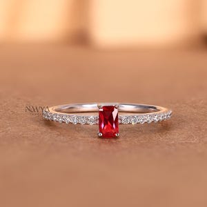 May include: A silver-toned ring featuring a central rectangular-cut red gemstone flanked by a row of small, clear, round-cut stones along the band. The word NAVYA is printed in small black text on the left side of the ring band.