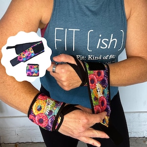 May include: Wrist wraps for weightlifting featuring a colourful donut pattern. The wraps are black with a vibrant design of donuts in various colours. The image also shows a matching wrist wrap and a small, square wrap with the same donut design.
