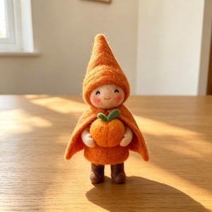 May include: A handmade felt doll in orange attire, holding an orange pumpkin with a green stem. The doll features a pointed hat and cape, with a smiling face. It stands on a wooden surface.