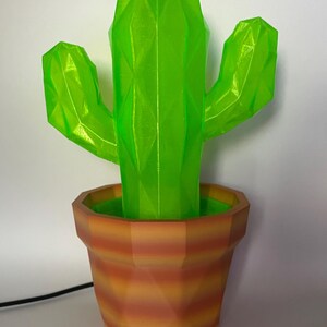 May include: A geometric, neon green cactus sculpture in a terracotta-colored pot. The cactus has a faceted design, with a central stalk and two side arms. The pot has a striped pattern, and a black cord extends from the base.