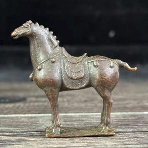 May include: A bronze-colored horse figurine with a saddle. The horse is standing on a rectangular base. The figurine has detailed features, including a mane, tail, and saddle. The horse is posed in a standing position, with its head slightly turned.
