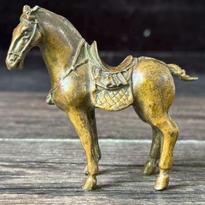 May include: A bronze-colored horse figurine with a saddle and bridle. The horse is posed in a standing position, with its head slightly turned. The figurine is detailed and appears to be made of metal.