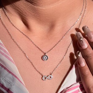 May include: A silver necklace with two chains. The top chain has a small, circular pendant with a blue and white pattern. The lower chain features an infinity symbol pendant, also silver. The necklace is worn over a pink and white striped top.