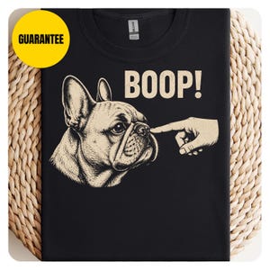 Peut inclure: Un t-shirt noir avec une illustration graphique beige d'un bouledogue français dont le museau est touché par un doigt. Le texte BOOP! est imprimé en beige au-dessus du chien. Un badge circulaire jaune avec le mot GUARANTEE est présent.