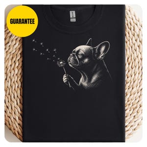 May include: A black t-shirt featuring a white graphic illustration of a French bulldog blowing on a dandelion. The dog is shown in profile, and the dandelion seeds are drifting away. A yellow circle with the text GUARANTEE is in the corner.