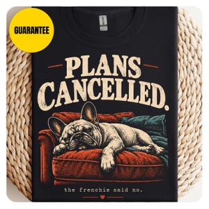 Peut inclure: Un t-shirt noir avec une illustration d'un bouledogue français dormant sur un canapé rouge avec un coussin bleu sarcelle. Le texte indique PLANS CANCELLED. et the frenchie said no. avec un petit cœur rouge au bas du motif.