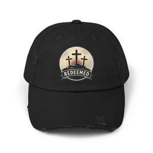 Puede incluir: Gorra de béisbol negra desgastada con un emblema circular que muestra tres cruces sobre una colina y la palabra "REDEEMED" debajo. El emblema está sobre un fondo beige y crema. La gorra tiene una visera curva y un aspecto desgastado.