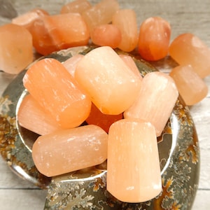 May include: A collection of peach-colored selenite crystals, some tumbled and others more natural, are arranged on a decorative, patterned dish. The crystals vary in size and shape, with a soft, translucent appearance. The background is a light-colored wooden surface.