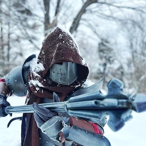 May include: A person in a medieval-style costume stands in a snowy environment. The costume includes a metal helmet with a cross, a brown hood, and metal armor. They hold a silver mace with a spiked head. The scene evokes a historical or fantasy theme.