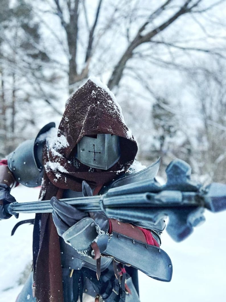 May include: A person in a medieval-style costume stands in a snowy environment. The costume includes a metal helmet with a cross, a brown hood, and metal armor. They hold a silver mace with a spiked head. The scene evokes a historical or fantasy theme.