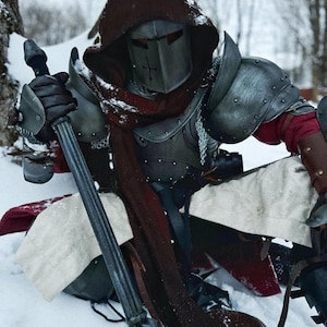 May include: A person in medieval armor, including a helmet with a cross, shoulder guards, and chainmail. The armor is a mix of silver and dark grey. The person is holding a long, dark weapon and is sitting in the snow. A red scarf is visible.