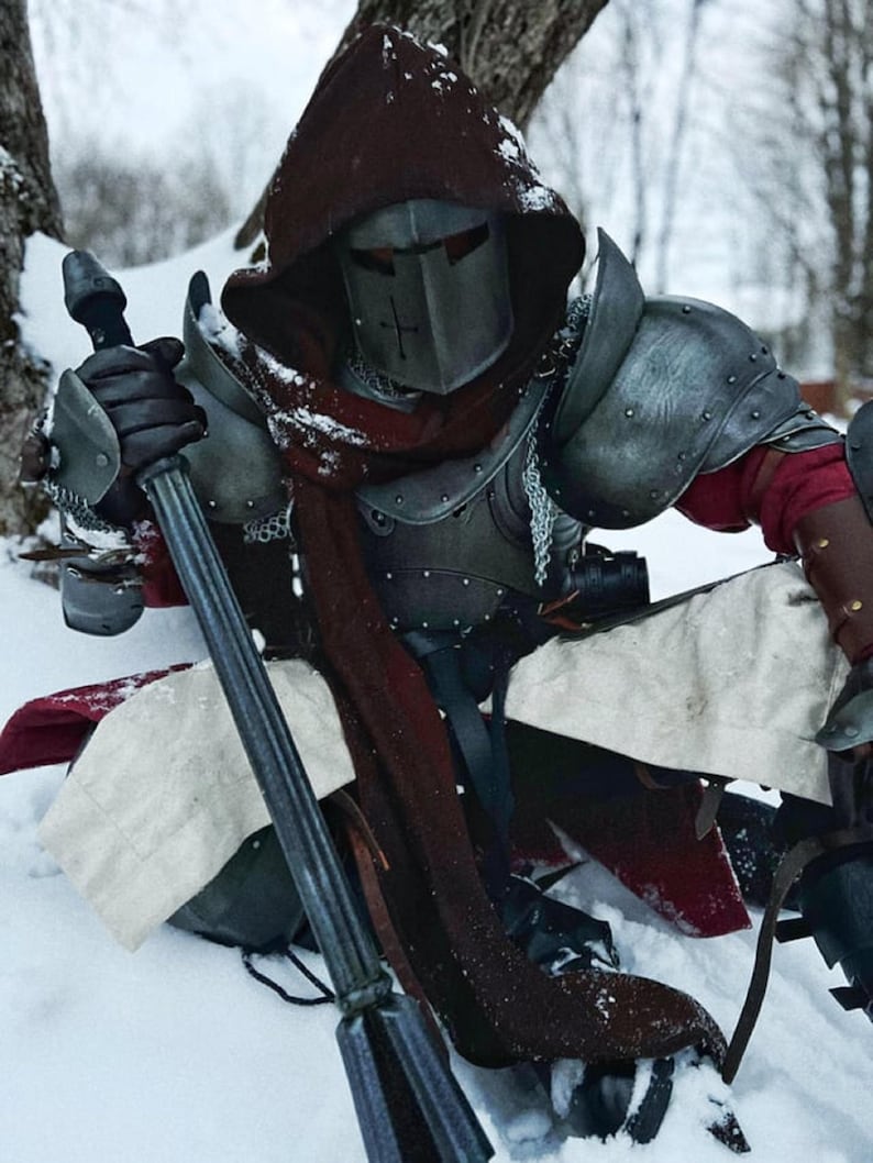 May include: A person in medieval armor, including a helmet with a cross, shoulder guards, and chainmail. The armor is a mix of silver and dark grey. The person is holding a long, dark weapon and is sitting in the snow. A red scarf is visible.