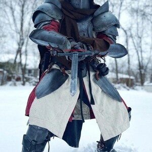 May include: A medieval knight in full armor stands in the snow, holding a sword. The armor is a mix of metal plates and leather, with a helmet and hood. The knight's attire includes a red undershirt and a white tunic with red trim.