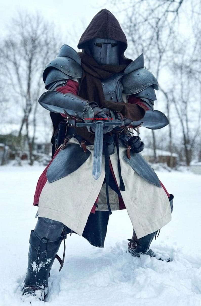 May include: A medieval knight in full armor stands in the snow, holding a sword. The armor is a mix of metal plates and leather, with a helmet and hood. The knight's attire includes a red undershirt and a white tunic with red trim.