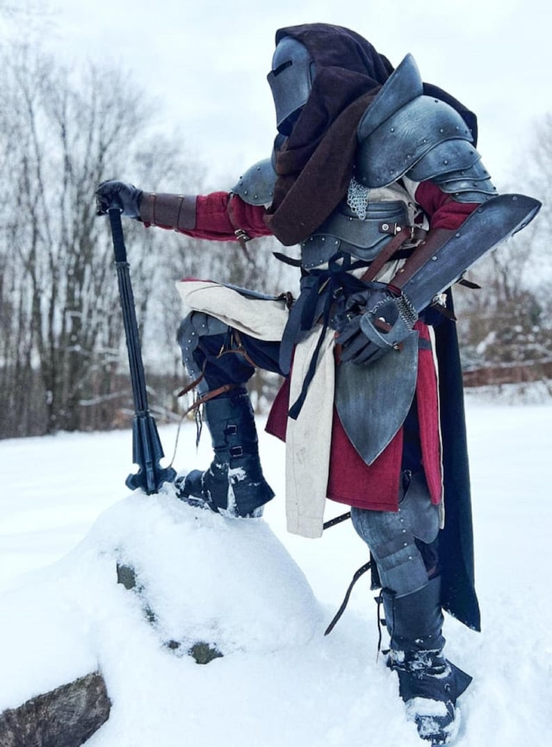 May include: A person in full medieval armor stands in the snow, holding a black polearm. The armor is a mix of metal and leather, with red and white fabric accents. The helmet has a visor, and a dark hood is worn.
