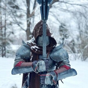 May include: A person in medieval armor holding a spiked mace. The armor is a weathered gray with red accents and chainmail. The person is wearing a brown hood and is standing in a snowy environment.