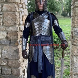 May include: A complete set of silver-coloured armour, including a helmet, chest plate, and leg guards. The armour is worn over a dark blue tunic and cape. A long sword is held in the right hand. The scene is set outdoors, framed by a stone archway.