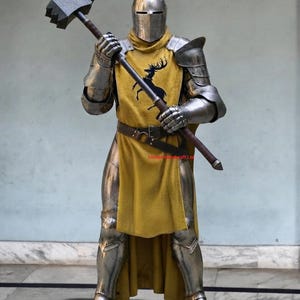 May include: A full suit of polished steel plate armour featuring a closed helmet, articulated gauntlets, and greaves. A mustard yellow tabard with a black stag emblem is worn over the armour, secured by a brown leather belt. A large steel war hammer is held.