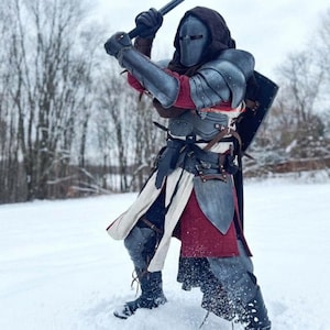 May include: A person in medieval-style armor, wielding a spiked mace in a snowy environment. The armor is a mix of silver and gray, with red and white accents. The person is wearing a helmet and a hooded cloak.
