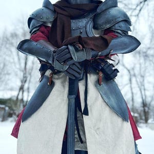 May include: A full suit of medieval armor, including a helmet with a cross design, shoulder guards, and arm protection. The armor is a dark, metallic color, and the person is wearing a brown hood and scarf. A long sword is held in front.