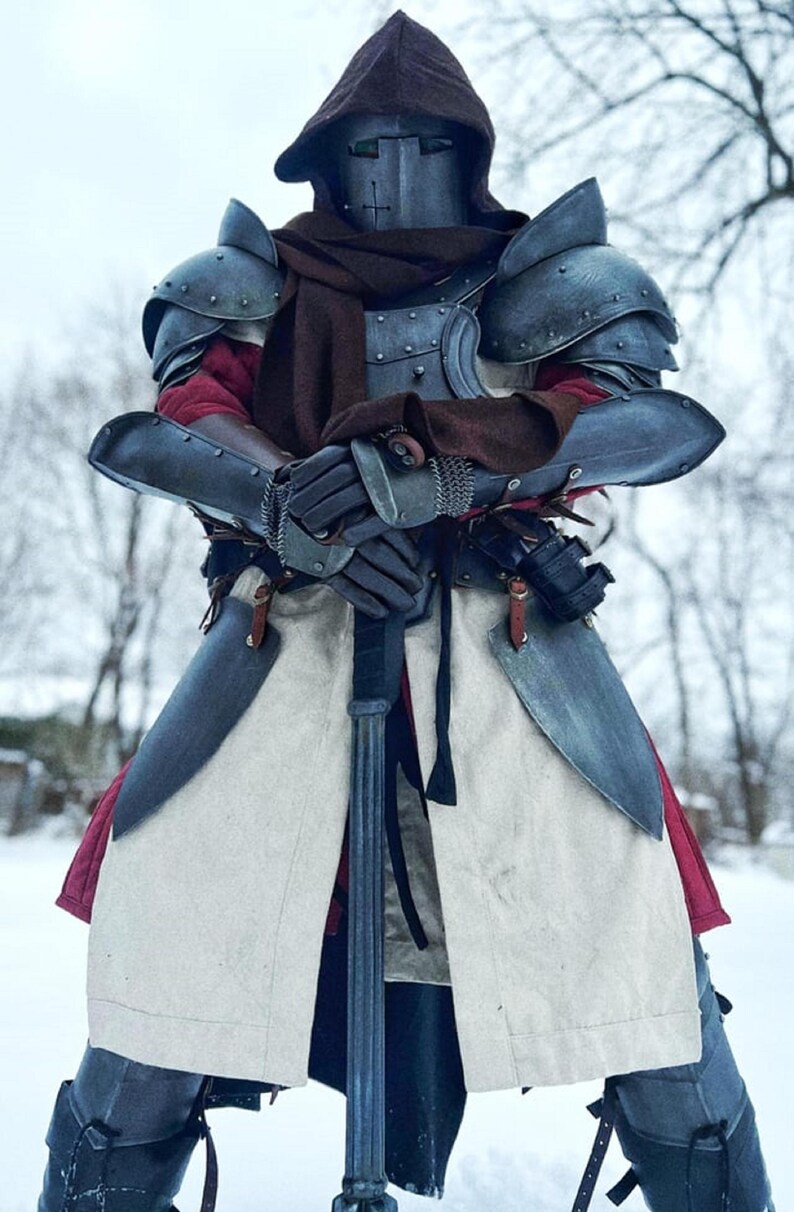 May include: A full suit of medieval armor, including a helmet with a cross design, shoulder guards, and arm protection. The armor is a dark, metallic color, and the person is wearing a brown hood and scarf. A long sword is held in front.