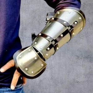 May include: A silver metal arm guard with rivets and brown leather straps. The armour covers the forearm and part of the hand, evoking a medieval or fantasy aesthetic. The measurements are in centimeters.