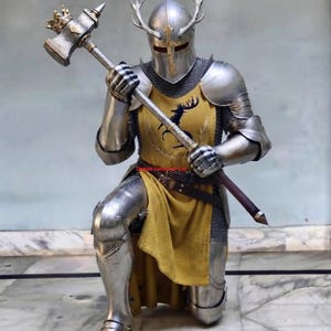 Puede incluir: Una armadura de placas de acero pulido que incluye un casco con astas y un detalle de cruz dorada. La armadura presenta un tabardo amarillo con un emblema de ciervo negro, capas de cota de malla y un gran martillo de guerra con corona.