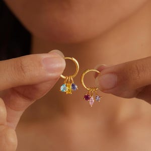 以下が含まれることがあります： 2つのゴールドフープイヤリング。それぞれにカラフルな宝石チャームが付いています。片方のイヤリングには青、黄、紫の宝石、もう片方にはピンクと紫の宝石が付いています。イヤリングはぼやけた背景に対して保持されています。