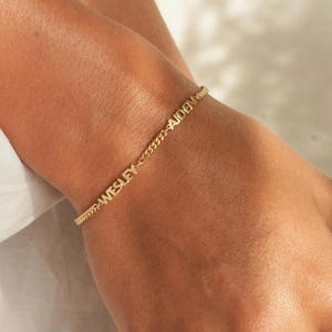 Puede incluir: Una pulsera de cadena dorada con los nombres "WESLEY" y "AIDEN" escritos en mayúsculas. La pulsera se lleva en la muñeca y la cadena tiene un diseño delicado y texturizado.