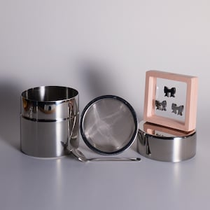 May include: Stainless steel tea canister with a mesh strainer, small spoon, and a decorative pink framed display with three black bow shapes. The items are arranged on a white surface.
