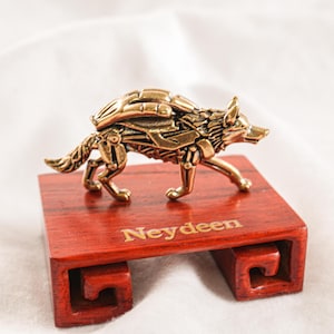 May include: A metallic brass-toned wolf figurine with mechanical, gear-like detailing across its body. The wolf stands atop a rectangular, reddish-brown wooden display base that features the gold-colored text Neydeen engraved on the front surface.