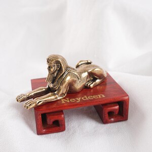 May include: A metallic bronze-toned sphinx figurine with detailed facial features and a headdress, resting on a rectangular reddish-brown wooden base. The word Neydeen is printed in gold lettering on the top surface of the wooden stand.