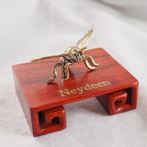May include: A metallic gold-toned insect figurine with detailed wings and legs, mounted on a rectangular reddish-brown wooden display base. The base features a carved scroll design on the sides and the word Neydeen printed in gold lettering.