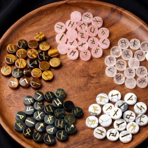 May include: A collection of circular stone discs arranged in five distinct groups on a wooden surface. Each disc features a gold-colored engraved runic symbol. The stones include polished tiger's eye, rose quartz, clear quartz, black obsidian, and white marble.