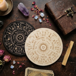 Puede incluir: Dos tableros de espiritismo circulares de madera, uno negro y otro de madera clara, con letras, números, meses, días, signos del zodiaco y una estrella central grabados. Los tableros están rodeados por un libro de cuero, cristales y pétalos de rosa.